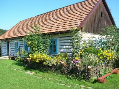 Drewniana chałupa od frontu otoczona kwiatami.