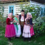 Trzy ubrane na ludowo kobiety stoją w ogrodzie, przez drewnianą chatą, między nimi mężczyzna w lachowskim stroju.
