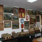 Ściana z wieloma obrazami, na długim stole makiety drewnianych domów.