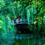 Mały staw z łukowym mostkiem zdobiony malowaniem w kwiaty, pośród bujnej zieleni.