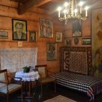 Wnętrze drewnianej izby, na ścianach wiszą obrazy, z prawej strony kolorowa makata.