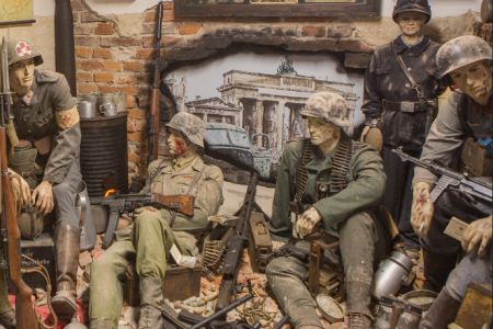Manekiny ubrane w wojskowe mundury, obok nich leżą karabiny, w tle grafika zniszczonego budynku.