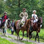 Osoby w strojach z epoki idą leśną ścieżką, dwie osoby jadą na koniach.