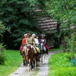 Grupa ludzi w dawnych strojach jedzie konno po ścieżce, w tle drzewa.