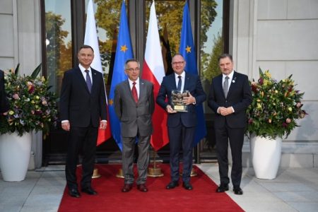 Czterech mężczyzn w garniturach stoi przed pałacem prezydenckim, jeden z nich trzyma statuetkę.