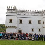 Zdjęcie grupowe osób stojących i kucających, w tle masywna budowla i drzewa.